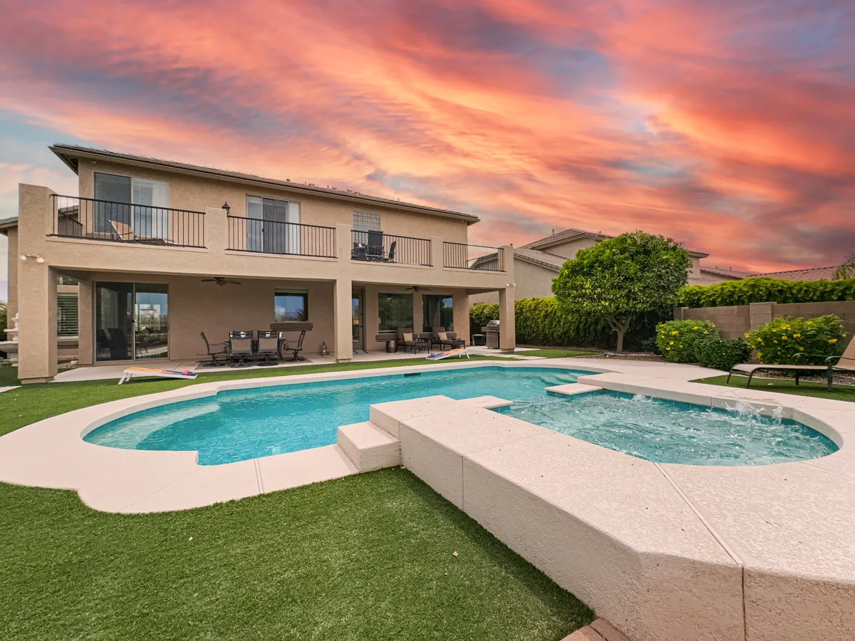 This exterior image features a sparkling swimming pool surrounded by a lush, landscaped garden, with a balcony overlooking the scene and a vivid sunset painting the sky. The pool area includes spots for outdoor dining and relaxing, making it perfect for group gatherings or tranquil evenings.