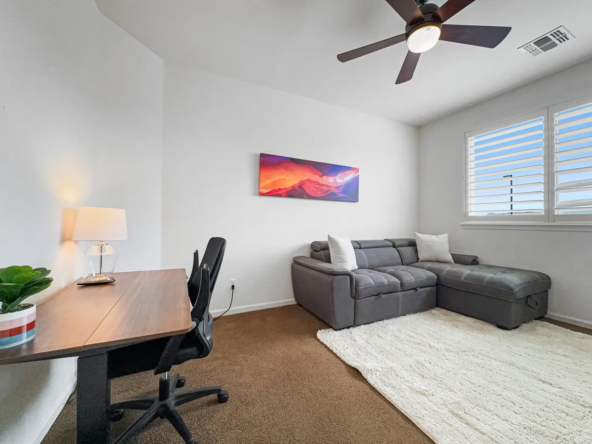 A multifunctional office/bedroom with a cozy gray sectional sofa, a practical wooden desk, colorful artwork on the wall, and a table lamp. The space is both inviting for relaxation and functional for focused work or reading.