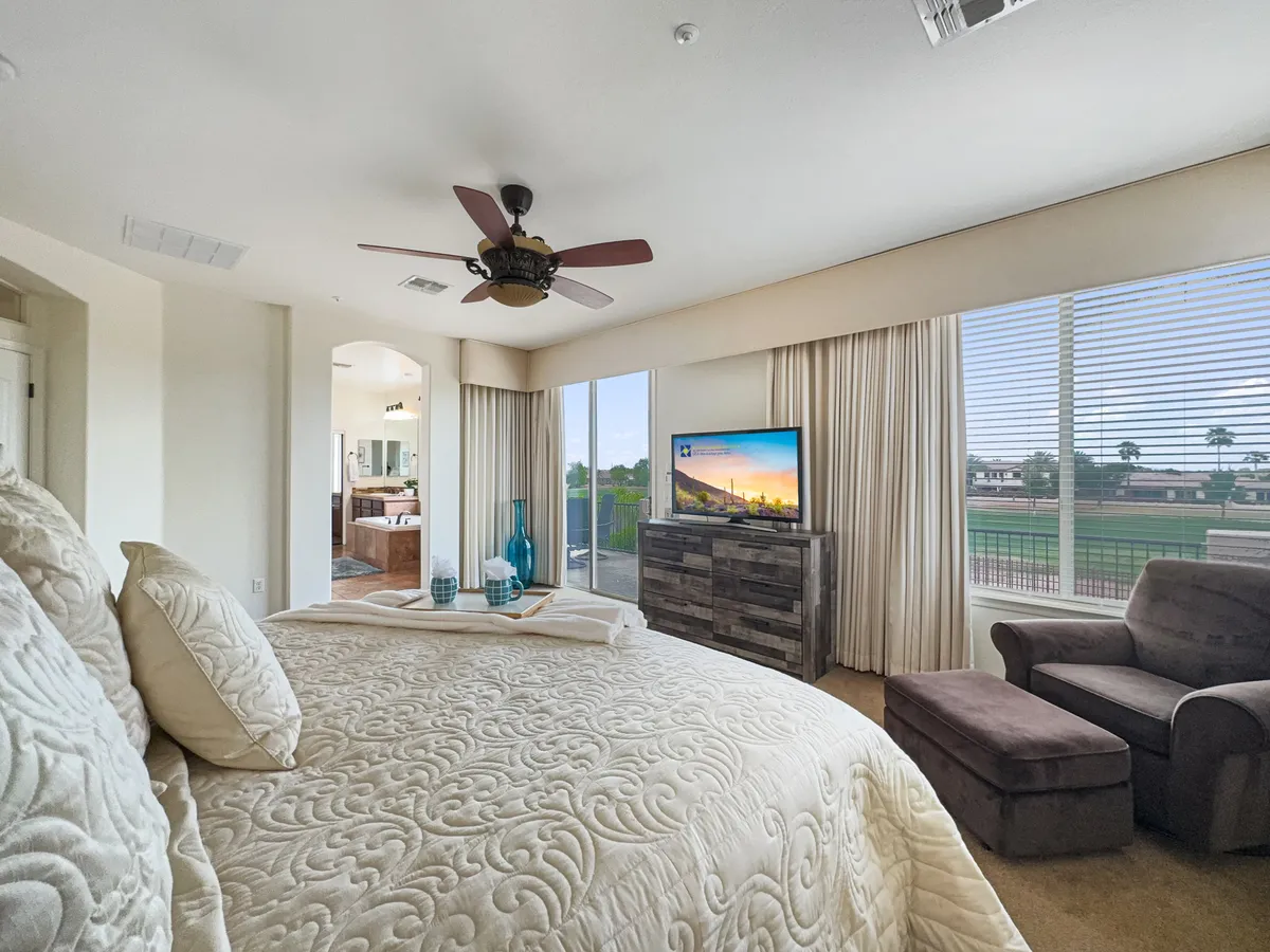 A serene bedroom featuring an intricately made bed with plush bedding, large windows adorned with flowing curtains that bathe the space in natural light, a wooden dresser, a comfortable armchair, and a wall-mounted television. The atmosphere is calming and ideal for unwinding.