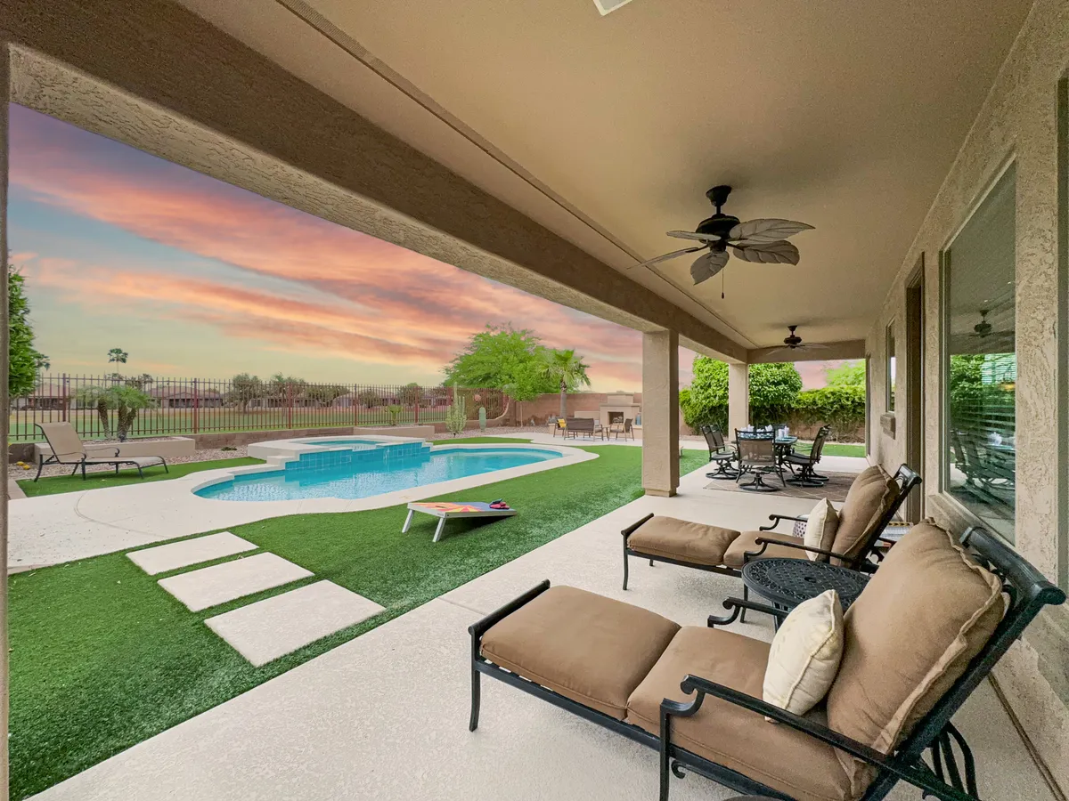 An inviting backyard scene with a shimmering pool, comfortable outdoor seating, lush landscaping, and a dramatic sunset sky. The patio area suggests a space ideal for entertaining guests or quiet reflection at any time of day.