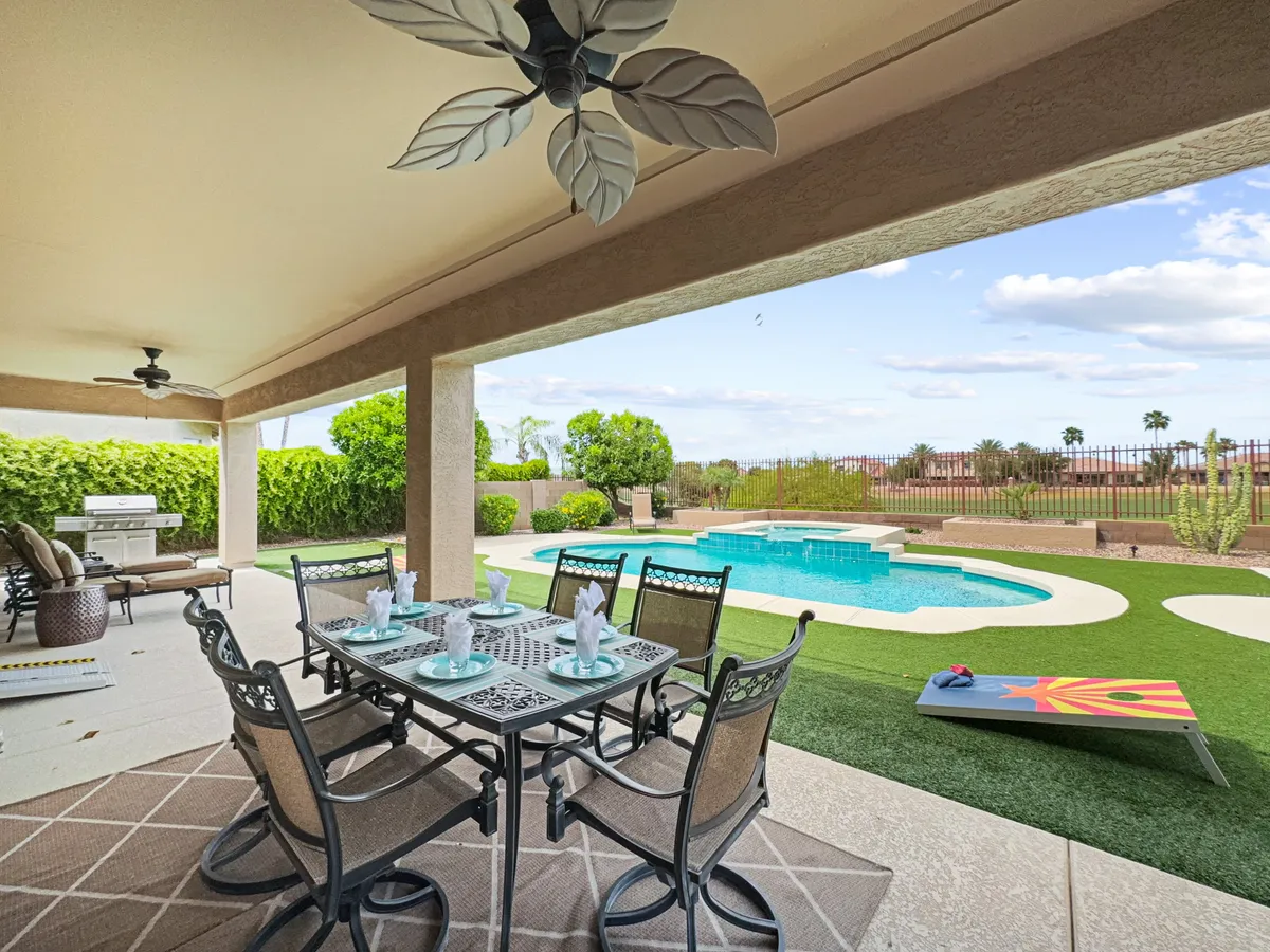 A lively backyard scene with a dining table and chairs set up for alfresco meals, a shimmering swimming pool, outdoor grill, and plenty of green lawn for games. The area is surrounded by a landscaped garden, making it ideal for both gatherings and playful activities.