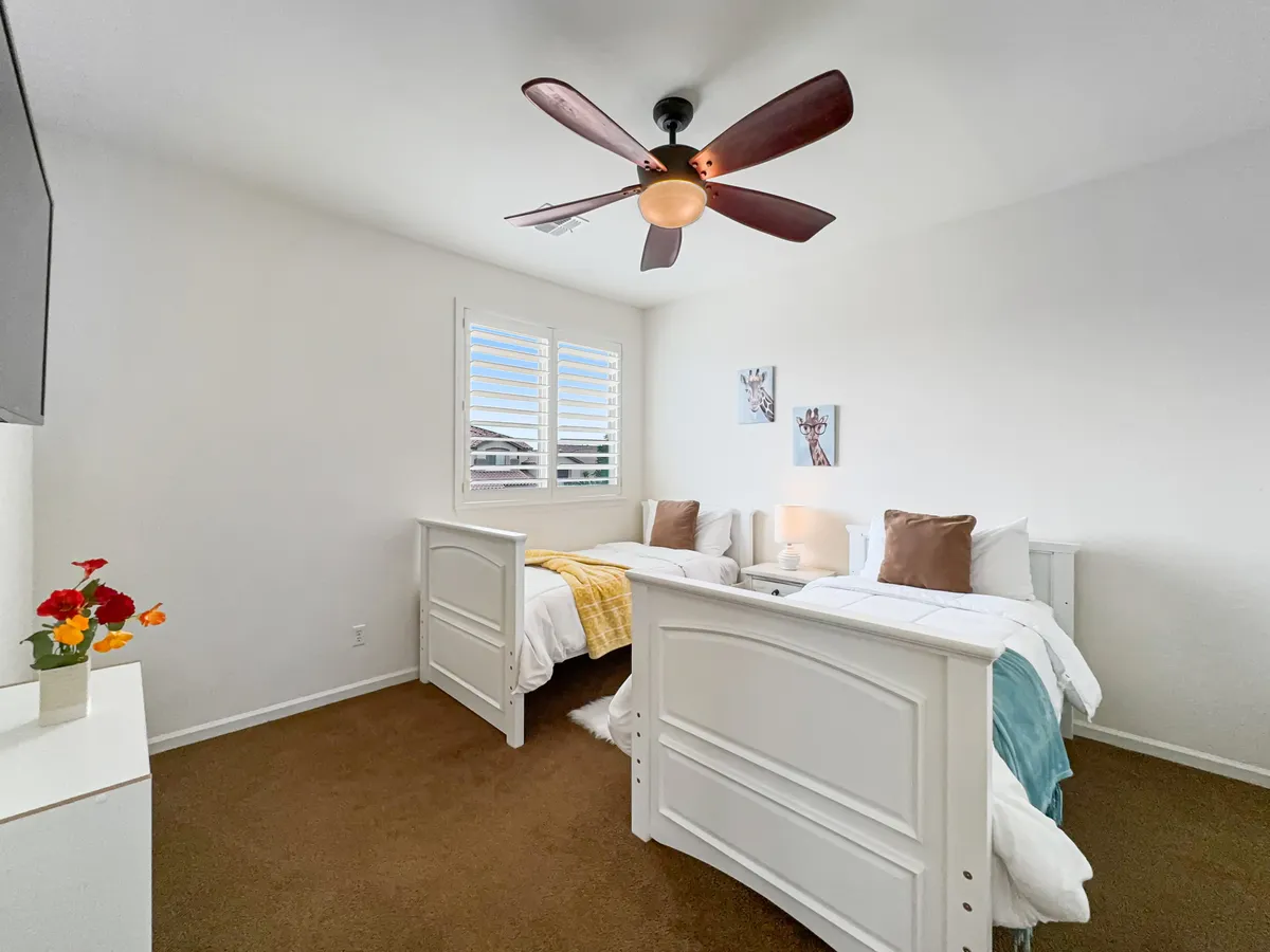 A bright and cheerful bedroom with two twin beds in white frames, vibrant flower arrangements, a decorative ceiling fan, and playful wall art. The room feels spacious and welcoming, ideal for guests or children.