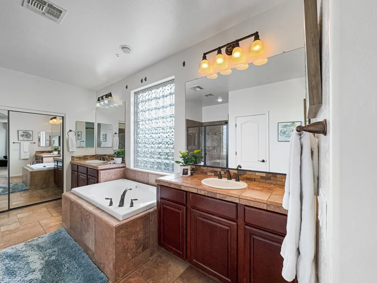 A tranquil spa-inspired bathroom with a luxurious soaking tub, double vanity, mirrors, towels, and a glass block window letting in soothing light. The space is elegant and designed for relaxation and self-care.